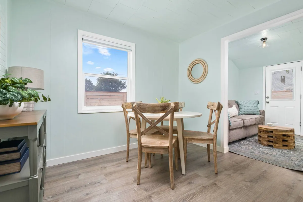 A close-up of the dining and living area with a focus on a stylish dining table, comfortable sofa, decorative mirror, and lush potted plant. The space feels cozy and light-filled, ideal for meals, reading, or socializing. The décor highlights the coastal theme with subtle, modern touches.