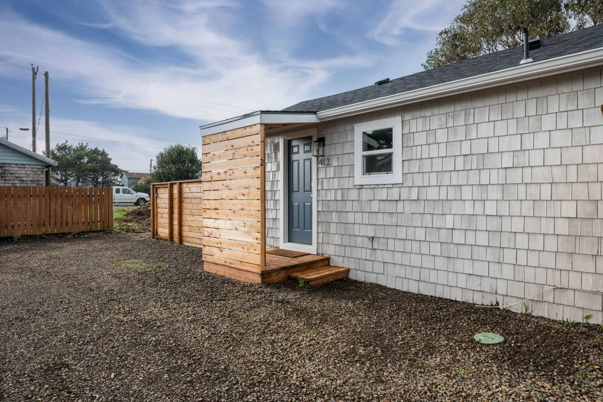 This image showcases the home's exterior front door, wooden steps, and a visible fenced yard. The inviting outdoor space is clearly defined, with the fencing providing a sense of privacy and security, matching the pet-friendly, enclosed nature of the yard. The welcoming steps and shingle siding reinforce a cozy, homey atmosphere.