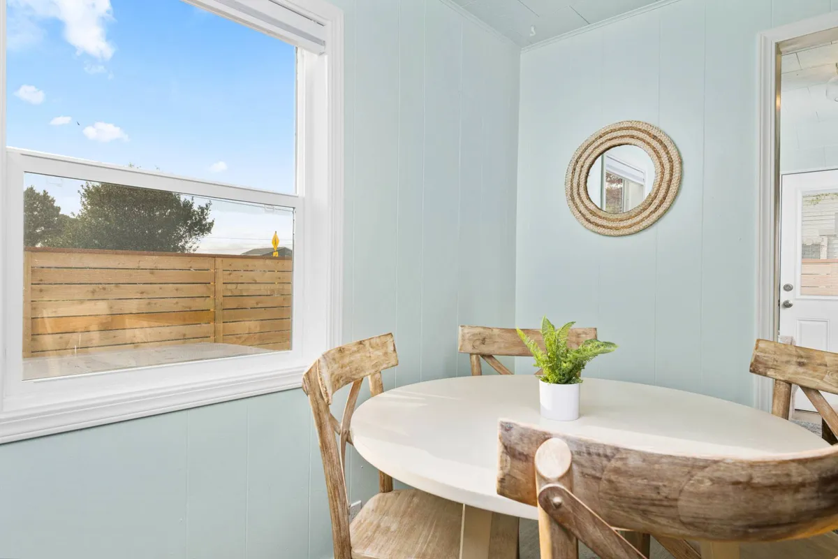 A bright and airy dining area with a round table surrounded by wooden chairs, a lush green plant, and a mirror reflecting natural light from the window. The view outside hints at the tranquil surroundings and the potential for indoor-outdoor living.