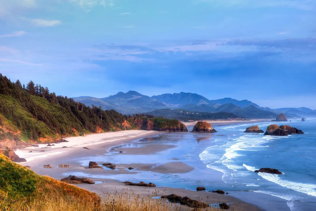 A tranquil shoreline stretches out with gentle ocean waves, rugged rock formations, and distant tree-lined mountains. The soft light hints at either early morning or sunset, with space for beachcombing, photography, and relaxed exploration.