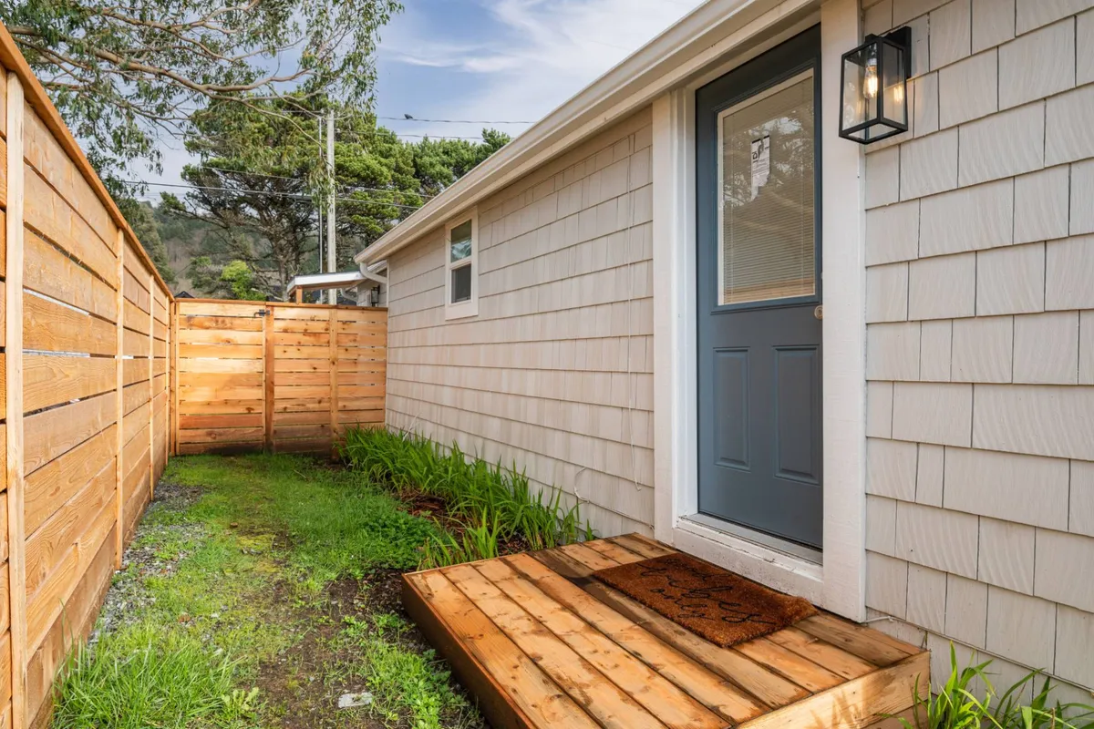 This image captures the exterior of the home with its inviting front door, wooden steps, and a fenced pathway leading to the backyard. The outdoor setting hints at private spaces beyond the entrance.