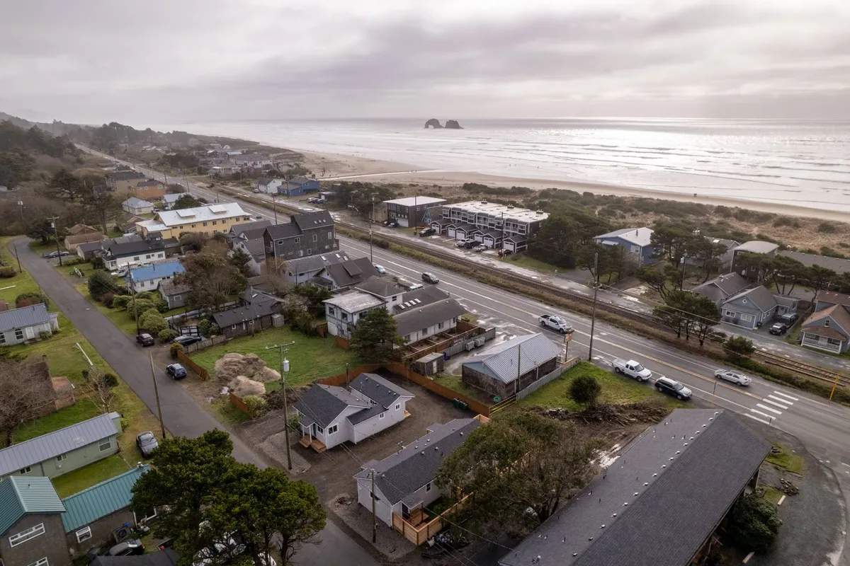A panoramic view featuring the ocean, sandy beach, nearby houses, and a road winding through the community. The image conveys a relaxed atmosphere, with people strolling on the beach, families picnicking, and hints of community gatherings or local dining spots close to the shoreline.