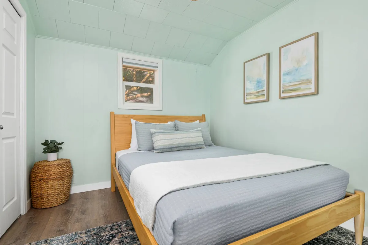 A tranquil bedroom featuring a neatly made full bed with inviting, plush linens. The décor is soft and calming, complemented by gentle wall art and touches of greenery from potted plants. Natural light filters in, creating a peaceful retreat ideal for rest and relaxation.